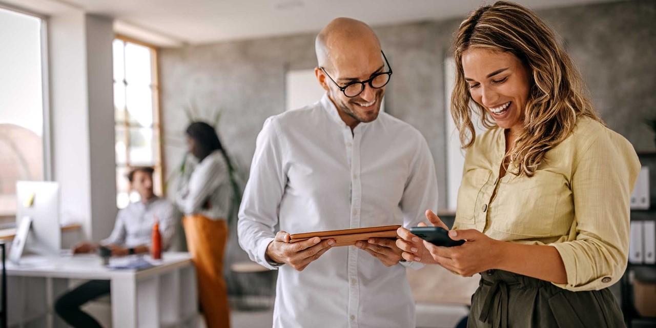 Cheerful and smiling young successful female businesswoman standing with colleague looking at smartphone in modern office and coworking space